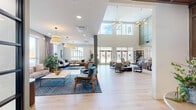 Located in Centennial, Colorado, this generously sized community clubhouse offers an open, single-floor layout designed for both comfort and functionality. The space makes an immediate impression with its soaring double-height ceilings in the central gathering area, anchored by a striking metal staircase with dark railings and flanked by tall paneled walls finished in soft sage green tones.

The expansive living area is thoughtfully furnished with multiple seating arrangements, including plush sectional sofas, leather lounge chairs, and patterned area rugs atop light-toned hardwood flooring. Large windows and floor-to-ceiling glass panels flood the space with natural light while offering views of the outdoor pool area, creating a bright and welcoming atmosphere throughout.

The kitchen is a standout feature, showcasing an extended white quartz island, sage green and warm wood-toned cabinetry with gold hardware, tile flooring, and a distinctive coffered wood ceiling overhead. The kitchen is fully appointed with stainless steel appliances and ample counter space, making it well-suited for community events and gatherings.

A clean, well-lit corridor lined with framed property signage connects additional rooms and utility spaces. The overall design palette — neutral walls, warm wood accents, and curated greenery — conveys a polished, modern aesthetic that is both inviting and functional.