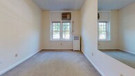 This single-floor unit is laid out in a straightforward, functional configuration that connects each area through a central hallway. The entry corridor features light ceramic tile flooring and white walls, leading from a warm wood-toned front door into the main living spaces. The kitchen is compact and efficiently arranged, equipped with warm oak cabinetry, white countertops, a four-burner electric range, an overhead ventilation hood, and a full-size refrigerator. Tile flooring continues throughout the kitchen and entry areas, providing a cohesive, easy-to-maintain surface.

The bedroom is carpeted and generously proportioned, benefiting from multiple windows that allow natural light to filter in. A wall-mounted mirror adds visual depth to the space. The room includes a window-unit air conditioner and a radiator, offering both heating and cooling options. Doors within the bedroom lead to additional spaces, including a closet area.

The bathroom is finished with classic square cream tiles lining the lower walls, complemented by white tile flooring. It includes a soaking bathtub with a shower fixture, a toilet, and a vanity. The unit's overall layout, as seen in the floorplan view, is elongated, with rooms arranged in a linear sequence that promotes a clear sense of flow throughout the space.