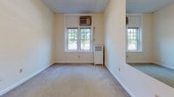 This single-floor unit is laid out in a straightforward, functional configuration that connects each area through a central hallway. The entry corridor features light ceramic tile flooring and white walls, leading from a warm wood-toned front door into the main living spaces. The kitchen is compact and efficiently arranged, equipped with warm oak cabinetry, white countertops, a four-burner electric range, an overhead ventilation hood, and a full-size refrigerator. Tile flooring continues throughout the kitchen and entry areas, providing a cohesive, easy-to-maintain surface.

The bedroom is carpeted and generously proportioned, benefiting from multiple windows that allow natural light to filter in. A wall-mounted mirror adds visual depth to the space. The room includes a window-unit air conditioner and a radiator, offering both heating and cooling options. Doors within the bedroom lead to additional spaces, including a closet area.

The bathroom is finished with classic square cream tiles lining the lower walls, complemented by white tile flooring. It includes a soaking bathtub with a shower fixture, a toilet, and a vanity. The unit's overall layout, as seen in the floorplan view, is elongated, with rooms arranged in a linear sequence that promotes a clear sense of flow throughout the space.