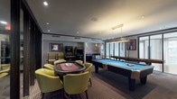 This single-level game room is thoughtfully designed for entertainment, offering a diverse array of recreational amenities within a stylish, modern interior. The space is anchored by a full-size pool table with a bright blue felt surface, positioned beneath a sleek gold pendant light fixture that provides focused illumination. Adjacent to the pool table, a round poker table surrounded by chartreuse upholstered chairs serves as a dedicated card-playing area, adding a lounge-like atmosphere to the room.

Along one wall, a pair of vintage arcade machines introduces a retro gaming element, complementing the room's eclectic character. A full-length shuffleboard table occupies a separate alcove, bordered by large floor-to-ceiling windows that flood the area with natural light and offer views of the surrounding exterior. A compact wet bar with dark granite countertops and a stainless steel mini-refrigerator provides convenient refreshment service within the space.

The room features recessed lighting throughout, warm neutral tones, and striped carpeting underfoot. Bold framed artwork and graphic canvas pieces decorate the walls. Black-framed glass partitions visually connect the game room to an adjoining space, enhancing the open, communal feel of the overall environment.