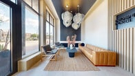This single-floor amenity space is thoughtfully designed to offer residents a well-rounded collection of communal features wrapped in a cohesive contemporary aesthetic. The welcoming lounge sets an impressive tone with soaring ceilings, floor-to-ceiling windows that flood the space with natural light, and sweeping views of the surrounding streetscape. A rich combination of materials — including dark slatted wall panels, polished concrete-look flooring, and a woven area rug — grounds the space, while sculptural pendant light fixtures serve as striking focal points overhead. Seating arrangements blend a tan leather sofa with warm wood-framed lounge chairs, creating an inviting and sophisticated atmosphere. An adjacent common area features a wood-clad island bar with sage green stools, wire pendant lighting, and wall-mounted screens, offering a versatile space for socializing or casual work. The fitness center is well-appointed with a wide range of cardio and strength equipment, including treadmills, stationary bikes, rowing machines, and free weights, all set on light hardwood flooring with large windows and floor-to-ceiling mirrors enhancing the bright, energetic environment. Completing the amenity suite is a dedicated dog spa, featuring tiled wet bath stations, a grooming area, and pet-friendly accessories — a thoughtful touch for four-legged residents.