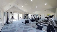 This fitness center offers a thoughtfully designed workout environment spread across a single floor. The space is defined by its striking vaulted ceiling, which adds a sense of openness and volume, complemented by recessed lighting and a ceiling fan that ensure a bright and comfortable atmosphere during exercise sessions. Large windows along one wall invite ample natural light, creating an energizing environment throughout the day. A full wall of floor-to-ceiling mirrors on the opposite side further enhances the sense of space while allowing users to monitor their form during workouts. The flooring features a modern geometric-patterned carpet in cool grey and blue tones, adding a contemporary aesthetic to the room. The facility is well-stocked with a variety of cardio equipment, including elliptical machines, stationary bikes, and treadmills, arranged to allow ease of movement throughout the space. Additional strength and multi-use training equipment is positioned toward the windowed wall, providing a well-rounded selection for all fitness levels. The overall ambiance is clean, bright, and functional, making this fitness center a welcoming and practical space for a comprehensive workout experience.