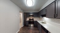 This single-floor unit offers a well-organized layout that balances functional living spaces with updated finishes throughout. The kitchen is a standout feature, showcasing dark charcoal cabinetry with sleek hardware, white subway tile backsplash, light countertops, and stainless steel appliances including a range, microwave, and dishwasher. Dark wood-style flooring grounds the kitchen, adding contrast to the lighter counters and walls.

The living room is distinguished by a floor-to-ceiling dark brick fireplace with a white mantle, providing a bold focal point. A sliding glass door with vertical blinds connects the space to an outdoor view of lush greenery, allowing natural light to filter in.

The bedrooms feature neutral-toned walls and plush carpeting, with windows overlooking outdoor greenery. One bedroom includes a double-door closet, offering practical storage. The bathroom maintains a clean, minimal aesthetic with white tile surrounding the tub and shower area, white fixtures, and a gold-toned towel bar accent.

Textured ceilings are consistent throughout the unit, and the overall color palette of warm grays and neutrals creates a cohesive, understated ambiance across all rooms.