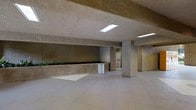 This single-floor space showcases a distinctly utilitarian architectural character, defined by its textured concrete surfaces that span both the walls and ceiling, giving the interior a raw, industrial aesthetic. The ceiling features recessed fluorescent lighting that provides consistent, even illumination throughout the open area. Large structural columns punctuate the floor plan, reinforcing the building's concrete construction while subtly dividing the space into functional zones. A notable built-in planter runs along one wall, adding a touch of greenery and softening the otherwise austere concrete palette. The flooring consists of large-format light-colored tiles laid in a grid pattern, offering a clean contrast to the rougher textures above. Toward the rear of the space, additional rooms or utility areas are visible, suggesting supplementary functional zones. The open layout offers considerable flexibility for a range of uses. Overall, the space communicates a no-frills, functional design sensibility with durable, low-maintenance materials throughout.