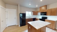 Located at 514 East Hastings Road in Spokane, WA, this single-floor unit offers a well-organized and practical layout designed for comfortable everyday living. The kitchen serves as a central highlight, featuring warm-toned wood cabinetry, a full suite of black appliances including a refrigerator, electric range, dishwasher, and over-the-range microwave. A center island with a light stone-style countertop provides additional prep space and casual seating potential. Light hardwood-style flooring runs throughout the kitchen, adding warmth and easy maintenance. The adjacent living area is generously sized, with light-toned walls, light flooring that reflects natural light from a window, and a secondary door providing exterior access. The bedroom is finished with neutral wall tones and soft carpeting underfoot, offering a comfortable and quiet retreat with a window for natural light. The bathroom includes a full tub and shower combination with a built-in shelf, a wood-toned vanity with a stone-style countertop, and a window for ventilation. Throughout the unit, neutral paint tones, recessed lighting in the kitchen, and consistent flooring choices create a cohesive, clean aesthetic that is move-in ready.