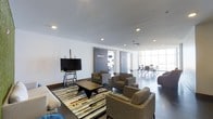 This single-floor clubroom and fitness center is designed with a clean, contemporary aesthetic that balances functionality with comfort. The main lounge area serves as the centerpiece, featuring a generous arrangement of plush upholstered armchairs and sofas grouped around a solid wood coffee table atop a bold, geometric area rug. A large flat-screen TV mounted on a mobile stand provides an entertainment focal point for the space. Recessed lighting throughout the smooth white ceiling creates a bright and welcoming ambiance. Sliding barn-style doors offer a sleek transition to the fitness area, which is visible through the opening and equipped with exercise equipment. Beyond the lounge, floor-to-ceiling windows flood the conference area with abundant natural light, where a meeting table and chairs are arranged for collaborative use. The polished dark flooring ties the entire space together, lending a sophisticated, professional feel. The open layout encourages movement between the lounge, fitness, and meeting zones, making this a versatile amenity space suited for relaxation, wellness, and productivity alike.