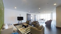 This single-floor clubroom and fitness center is designed with a clean, contemporary aesthetic that balances functionality with comfort. The main lounge area serves as the centerpiece, featuring a generous arrangement of plush upholstered armchairs and sofas grouped around a solid wood coffee table atop a bold, geometric area rug. A large flat-screen TV mounted on a mobile stand provides an entertainment focal point for the space. Recessed lighting throughout the smooth white ceiling creates a bright and welcoming ambiance. Sliding barn-style doors offer a sleek transition to the fitness area, which is visible through the opening and equipped with exercise equipment. Beyond the lounge, floor-to-ceiling windows flood the conference area with abundant natural light, where a meeting table and chairs are arranged for collaborative use. The polished dark flooring ties the entire space together, lending a sophisticated, professional feel. The open layout encourages movement between the lounge, fitness, and meeting zones, making this a versatile amenity space suited for relaxation, wellness, and productivity alike.