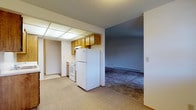 This single-floor residential unit offers a practical and straightforward layout designed for comfortable everyday living. The galley-style kitchen is efficiently organized with warm wood cabinetry lining both sides, white appliances including an electric range and refrigerator, a dishwasher, and a patterned vinyl floor. A drop ceiling with recessed panel lighting brightens the kitchen area. The kitchen opens conveniently to the main living space, which features neutral wall-to-wall carpeting, a textured ceiling, and a baseboard heating system. A sliding glass door with vertical blinds floods the living area with natural light and provides direct access to the outdoors. The bedroom is a generously sized, carpeted room with its own window fitted with blinds, a textured ceiling, and baseboard heating, offering a quiet and comfortable retreat. A secondary carpeted room serves as additional flexible living space, connected to the kitchen and hallway. The full bathroom is clean and functional, featuring a wood-toned vanity with a single sink and drawers, a large mirror, a tub-shower combination, and neutral tile flooring. Throughout the unit, the design is practical and understated, with light-toned walls creating a bright and open atmosphere.