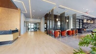 This contemporary hospitality space makes a striking first impression with its expansive, open-plan layout spanning a single thoughtfully designed floor. The grand entrance foyer welcomes guests with light wood-plank flooring, soaring ceilings embedded with warm linear LED lighting, and floor-to-ceiling glass doors that flood the space with abundant natural light and offer views of the surrounding streetscape.

At the heart of the space lies a dramatic lounge area framed by dark, veined marble accent walls and cascading metallic chain curtains that serve as elegant room dividers. Bold red velvet seating, cordoned off with gold stanchions, creates an exclusive VIP atmosphere. Transitioning into the bar area, guests are greeted by a sleek, dark marble-topped bar counter lined with stylish blue and amber upholstered barstools. A striking geometric shelving unit displays curated bar offerings behind an industrial-style black metal frame.

The dining area benefits from floor-to-ceiling windows along the street-facing facade, providing ample natural light. Geometric patterned tilework on both walls and floors adds visual depth and character throughout. A dedicated reception desk, clad in dark marble with brass accents, anchors one section of the space, complemented by textured bronze wall panels. The overall ambiance is refined, modern, and unmistakably upscale.