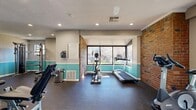This fitness center offers a comprehensive workout environment thoughtfully designed to accommodate a wide range of exercise needs. The space features a distinctive two-tone wall treatment, with a warm beige upper half and a bold teal lower half accented by white chair rail molding, lending the room a clean and energetic aesthetic. Exposed brick walls add a touch of industrial character, particularly in the cardio zone, where large picture windows flood the area with natural light and provide views of the surrounding outdoors.

The cardio section is equipped with a treadmill, elliptical trainer, and additional aerobic machines, all positioned to take advantage of the natural light and outdoor views. A dedicated strength training area houses multiple cable and resistance machines, a multi-function cable tower, adjustable benches, and a full rack of dumbbells in varying weights, catering to users of all fitness levels. A wall-mounted flat-screen television is positioned for convenient viewing during workouts.

Durable rubber flooring throughout provides safety and comfort underfoot. Recessed ceiling lighting ensures consistent illumination across the entire space. Entry is provided through elegant white French doors with glass panes, which also enhance the flow of natural light. Fitness center rules are posted on the wall, and the space is well-ventilated with visible HVAC fixtures.