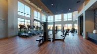 This expansive fitness center is designed to offer a premium workout experience within a bright, open, and architecturally striking environment. The space is defined by soaring double-height ceilings and an extensive array of floor-to-ceiling windows that wrap around the facility, flooding the interior with abundant natural light and offering unobstructed views of the surrounding outdoor landscape. Rich hardwood-style flooring runs throughout, adding warmth and a polished aesthetic that complements the modern design. The cardio zone is generously equipped with multiple treadmills, elliptical machines, and upright bikes, all positioned to take full advantage of the panoramic window views. A dedicated strength training area features a comprehensive free weight section with a full rack of dumbbells, benches, and large wall mirrors that enhance the sense of space. Functional training is well-represented with a dual cable cross machine, a multi-station strength unit, resistance bands, medicine balls, kettlebells, and stability equipment neatly organized on racks. Recessed lighting and a tray ceiling detail in one section of the space add visual depth and architectural interest. The overall ambiance is energetic yet refined, making this fitness center a standout amenity that caters to a wide range of workout preferences and fitness levels.