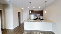 This single-floor residence showcases a thoughtfully designed open-concept layout that seamlessly connects the kitchen and living areas. The kitchen is a standout feature, highlighted by rich dark wood cabinetry that provides generous storage and a striking visual contrast against the light-toned granite countertops. A spacious peninsula extends the counter space, offering a functional area for dining or entertaining. Stainless steel appliances, including a refrigerator, microwave, and range, lend a sleek, contemporary feel to the space. Recessed lighting and elegant pendant lights suspended above the peninsula ensure the kitchen is well-illuminated, creating a warm and inviting atmosphere. The flooring throughout is a deep, rich hardwood-style material that adds warmth and continuity across the open floor plan. Neutral-toned walls complement the overall palette, keeping the space feeling bright and airy. A glimpse into the adjoining hallway reveals additional rooms, including what appears to be a walk-in closet, hinting at the thoughtful storage solutions integrated throughout the unit. The clean lines, quality finishes, and cohesive design make this a polished and practical living space.