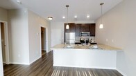 This single-floor residence showcases a thoughtfully designed open-concept layout that seamlessly connects the kitchen and living areas. The kitchen is a standout feature, highlighted by rich dark wood cabinetry that provides generous storage and a striking visual contrast against the light-toned granite countertops. A spacious peninsula extends the counter space, offering a functional area for dining or entertaining. Stainless steel appliances, including a refrigerator, microwave, and range, lend a sleek, contemporary feel to the space. Recessed lighting and elegant pendant lights suspended above the peninsula ensure the kitchen is well-illuminated, creating a warm and inviting atmosphere. The flooring throughout is a deep, rich hardwood-style material that adds warmth and continuity across the open floor plan. Neutral-toned walls complement the overall palette, keeping the space feeling bright and airy. A glimpse into the adjoining hallway reveals additional rooms, including what appears to be a walk-in closet, hinting at the thoughtful storage solutions integrated throughout the unit. The clean lines, quality finishes, and cohesive design make this a polished and practical living space.