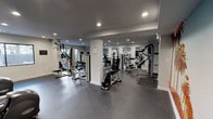 This fitness center is a comprehensively equipped, single-floor facility designed to support a full range of workout routines. The space is divided into distinct functional zones, seamlessly connected to create an open and accessible training environment.

The cardio area features multiple treadmills, elliptical trainers, and a stationary bike, all positioned beneath ceiling-mounted televisions for entertainment during workouts. A bold, eye-catching mural depicting a sunny palm tree boulevard serves as a striking accent wall, adding a vibrant, energetic atmosphere to this section of the facility.

The strength training zone offers a variety of cable machines, multi-functional weight stations, and benches, providing options for both upper and lower body conditioning. Equipment is arranged to maximize floor space and ease of movement between stations.

Throughout the facility, large mirrors line the walls, enhancing the sense of spaciousness and allowing users to monitor their form. Recessed lighting illuminates the entire space evenly, while windows along the perimeter bring in natural light and views of the surrounding outdoors. The durable, rubberized dark flooring provides cushioning and stability underfoot.

The overall design is clean and modern, with a neutral color palette of white walls and grey tones that keeps the focus on the equipment and the workout experience.