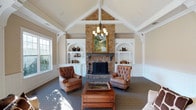 This single-floor space makes a striking impression with its centerpiece great room, where a soaring vaulted ceiling with exposed white timber accents draws the eye upward. A grand floor-to-ceiling stone fireplace anchors the room, flanked symmetrically by built-in white shelving units with arched detailing — ideal for displaying books and décor. The warm, neutral wall tones complement the natural stone and rich woodwork throughout, creating a cohesive and inviting aesthetic. A classic chandelier hangs from the peak of the vault, casting a warm glow over the seating area below. Tufted leather armchairs and a wooden coffee table with drawer storage furnish the space in a timeless, traditional style. Large windows on either side of the room allow ample natural light to pour in, highlighting the textured stone and crisp white millwork. Wainscoting along the lower walls adds architectural depth and a refined character consistent with the room's overall craftsmanship. The space exudes a warm, welcoming ambiance well-suited for relaxed gathering or quiet enjoyment, combining classic architectural details with comfortable, purposeful furnishings in a thoughtfully designed layout.