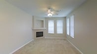 This single-floor space features a spacious living area finished in a neutral, light beige palette that creates a clean and welcoming atmosphere. The room is anchored by a built-in fireplace framed with a structured mantel surround and a recessed niche above, providing a natural focal point and an ideal spot for a display or media setup. Soft, wall-to-wall carpeting in a warm sand tone covers the floor, adding comfort underfoot and contributing to the room's understated, relaxed aesthetic. Overhead, a ceiling fan with an integrated light fixture provides both air circulation and ambient lighting. Natural light fills the room through multiple windows dressed with white horizontal blinds, offering flexibility in light control while maintaining a bright and airy feel. The walls are smooth and painted in a consistent light neutral tone, complementing the carpeting and overall design. Wide baseboards add a subtle architectural detail throughout. The open layout of the living space allows for versatile furniture arrangements, making it well-suited for a variety of uses. The overall ambiance is calm, clean, and move-in ready.