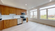 This contemporary single-floor unit showcases a thoughtfully designed open-concept layout that seamlessly connects the kitchen and living area. The kitchen is a standout feature, outfitted with rich warm-toned wood cabinetry that extends floor to ceiling, a white diamond-pattern tile backsplash, light quartz countertops, and a full suite of stainless steel appliances including a range, over-the-range microwave, and dishwasher. The light-toned hardwood flooring flows continuously throughout the space, enhancing the sense of openness and warmth. Large, multi-pane windows in the living area flood the space with natural light and frame elevated views of the surrounding cityscape and treetops, adding a dynamic urban backdrop to the interior. The bedroom is a calm, neutral retreat featuring the same light hardwood flooring, a ceiling fan, and a mirrored sliding closet door that reflects light and adds depth to the room. The bathroom is finished in a clean, minimalist style with full-height white rectangular wall tiles in the tub surround, a floating vanity with a wood-toned base and integrated white sink, a large mirror, and neutral floor tiles. Together, the finishes and layout create a cohesive, modern living environment.