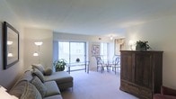 This single-floor residence offers a well-appointed and open living area that immediately conveys a sense of comfort and functionality. The living room is generously sized, featuring plush wall-to-wall carpeting that adds warmth underfoot and a soft, neutral palette throughout. Large sliding glass doors span a significant portion of one wall, flooding the interior with natural light and offering views of the surrounding greenery outside, creating a seamless connection between the indoors and the exterior environment. The open layout accommodates both a relaxed seating area, anchored by a spacious sectional sofa, and a dedicated dining nook furnished with elegant wrought-iron chairs and a glass-top table — an ideal arrangement for everyday living and casual entertaining. A substantial dark-wood armoire adds a rich, classic contrast to the lighter tones of the room and provides ample storage. Tasteful artwork adorns the walls, contributing to the refined yet approachable ambiance of the space. Ceiling-mounted and floor lighting fixtures ensure the interior remains well-lit throughout the day and into the evening. The overall atmosphere is calm, airy, and inviting, making this a comfortable and practical space suited for a variety of lifestyles.
