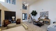 This refined single-floor space in Fort Worth presents an impressive and welcoming interior defined by its dramatic double-height ceilings that immediately establish a sense of openness and grandeur. A tasteful chandelier anchors the main living area, casting warm light across the neutral-toned carpeted floors and crisp white walls. The space is thoughtfully furnished with a curated mix of seating options, including a rich brown leather armchair, modern accent chairs, and a substantial dark-toned coffee table that serves as a focal point for the central gathering area. Large floor-to-ceiling windows and glass-paneled entry doors allow generous natural light to pour in, enhancing the bright and airy ambiance throughout. A statement fiddle-leaf fig tree adds a lush, organic touch that contrasts beautifully with the clean architectural lines. Decorative artwork and carefully arranged accessories on a warm wood console table contribute to the space's polished, contemporary aesthetic. The overall atmosphere is calm, inviting, and professionally appointed, making it well-suited for both relaxed social interaction and more formal engagements.