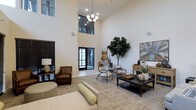 This refined single-floor space in Fort Worth presents an impressive and welcoming interior defined by its dramatic double-height ceilings that immediately establish a sense of openness and grandeur. A tasteful chandelier anchors the main living area, casting warm light across the neutral-toned carpeted floors and crisp white walls. The space is thoughtfully furnished with a curated mix of seating options, including a rich brown leather armchair, modern accent chairs, and a substantial dark-toned coffee table that serves as a focal point for the central gathering area. Large floor-to-ceiling windows and glass-paneled entry doors allow generous natural light to pour in, enhancing the bright and airy ambiance throughout. A statement fiddle-leaf fig tree adds a lush, organic touch that contrasts beautifully with the clean architectural lines. Decorative artwork and carefully arranged accessories on a warm wood console table contribute to the space's polished, contemporary aesthetic. The overall atmosphere is calm, inviting, and professionally appointed, making it well-suited for both relaxed social interaction and more formal engagements.