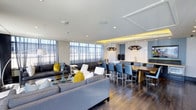 This single-floor space showcases a thoughtfully designed open-concept layout that seamlessly blends a generous lounge area with a formal dining space. The living area is anchored by plush grey sofas accented with bold yellow and white throw pillows, arranged around a low coffee table adorned with elegant decorative vases. Dark hardwood flooring runs throughout, providing a rich contrast to the light-toned walls and bright white ceiling.

Floor-to-ceiling windows dressed in sheer curtains line one wall, allowing abundant natural light to permeate the space and creating an airy, welcoming atmosphere. The dining area accommodates a large rectangular wood table surrounded by blue upholstered chairs, positioned beneath striking sculptural pendant lights that serve as a focal point.

A built-in wet bar or kitchenette anchors the far end of the room, featuring clean cabinetry and countertops. Cove lighting along a tray ceiling detail adds warmth and visual depth to the dining zone. The overall design palette of greys, blues, and yellow accents creates a modern, sophisticated ambiance suited for both relaxed gatherings and formal entertaining.
