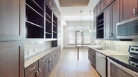 This single-floor space showcases a cohesive and modern design centered around a generously proportioned kitchen. Rich, dark-stained shaker-style cabinetry lines both sides of the kitchen, extending floor to ceiling to maximize storage capacity. The cabinetry is complemented by light granite countertops and a subtle glass tile backsplash, creating an elegant contrast throughout the space. A full suite of stainless steel appliances — including a range with electric cooktop, built-in microwave, dishwasher, and a French door refrigerator — provides a sleek, contemporary finish. Pendant lighting hangs above a built-in dining table at the kitchen's end, where large windows fitted with white blinds and transom windows above allow natural light to fill the area. Open shelving units on one wall add a decorative and functional display element. Light hardwood flooring flows seamlessly from the kitchen into the adjacent open living area, which features a tray ceiling detail that adds architectural interest. The bathroom is well-finished with large-format tile flooring, a granite-topped dark wood vanity, wall sconces, and a full tub-shower combination with clean, floor-to-ceiling tile surround. The overall ambiance is modern and refined.
