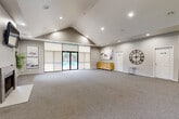 Located at 8211 S 87th Plaza in La Vista, Nebraska, this single-floor multipurpose community space offers a generously sized, open-plan layout ideal for gatherings and events. The room's most striking architectural feature is its vaulted ceiling, which soars to a dramatic peak and creates an impressive sense of height and volume throughout the space.

Natural light floods the interior through a wall of large sliding glass doors fitted with roller shades, offering direct views and access to an outdoor pool area. Additional windows on the adjacent wall further brighten the interior. Recessed lighting is installed throughout the ceiling, providing consistent, even illumination across the entire floor area.

The flooring is covered with neutral-toned, textured carpet that complements the soft gray walls and white trim, contributing to a clean and modern aesthetic. A built-in fireplace anchors one end of the room, adding a welcoming focal point. Multiple white panel doors lead to adjoining spaces, including what appears to be a catering kitchen, as well as restroom facilities.

Decorative touches including framed artwork, a large statement wall clock, and warm wood-toned storage dressers add character without compromising the room's versatile, open feel. The overall ambiance is contemporary, bright, and well-appointed — well-suited for community events, social gatherings, or private functions.