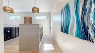 Located in New Port Richey, Florida, this well-appointed community mailroom and hallway space offers a clean, contemporary design with a coastal aesthetic. The open layout accommodates multiple freestanding mailbox islands clad in crisp white shiplap paneling, each housing rows of sleek, dark-finished individual mail compartments — providing convenient mail access for a large residential community.

Light wood-tone flooring runs throughout, complementing the bright white walls and contributing to an airy, welcoming atmosphere. Natural light filters in through strategically placed horizontal clerestory windows, while recessed ceiling lighting and distinctive woven pendant light fixtures add warmth and visual interest overhead.

The hallway leading away from the main mailroom area is equally refined, featuring the same light hardwood flooring, tall white walls, and clean-lined doorways with matte black hardware. A large-scale abstract artwork in rich oceanic blues and teals commands attention as a focal point on the main wall, setting a vibrant coastal tone throughout the space.

Decorative touches — including circular woven wall art and additional coastal-inspired paintings — reinforce the cohesive design theme. The space flows logically, with clear sightlines and an uncluttered layout that ensures ease of navigation. Overall, this mailroom and hallway space strikes a balance between practical functionality and thoughtful, upscale interior design.