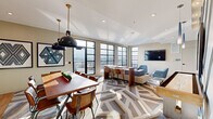 Located at 600 West State Street in Kennett Square, PA, this thoughtfully designed lounge space offers a seamless blend of modern aesthetics and functional comfort across a single floor. The open-concept layout unifies the living room, dining room, and hallway into one cohesive environment anchored by a striking geometric-patterned area rug that flows throughout the interior.

The living area is bathed in natural light, courtesy of expansive floor-to-ceiling steel-framed windows that frame panoramic outdoor views. Plush sectional seating and blue velvet accent chairs are arranged around a sleek coffee table, creating an inviting social atmosphere. A wall-mounted flat-screen television completes the lounge setting.

The dining area features a warm wood communal table with mixed seating and is illuminated by bold industrial pendant lights. Adjacent to it, a well-appointed kitchenette showcases a vibrant cobalt blue tile backsplash, light wood cabinetry, open shelving, and stainless steel appliances — adding both function and visual interest.

A standout feature is the bold teal accent wall with a diagonal wood-panel design, serving as a dramatic backdrop to a shuffleboard table — a playful and social focal point. The space extends outdoors to a tiled terrace with seating, offering an open-air retreat under natural light.