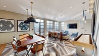 Located at 600 West State Street in Kennett Square, PA, this thoughtfully designed lounge space offers a seamless blend of modern aesthetics and functional comfort across a single floor. The open-concept layout unifies the living room, dining room, and hallway into one cohesive environment anchored by a striking geometric-patterned area rug that flows throughout the interior.

The living area is bathed in natural light, courtesy of expansive floor-to-ceiling steel-framed windows that frame panoramic outdoor views. Plush sectional seating and blue velvet accent chairs are arranged around a sleek coffee table, creating an inviting social atmosphere. A wall-mounted flat-screen television completes the lounge setting.

The dining area features a warm wood communal table with mixed seating and is illuminated by bold industrial pendant lights. Adjacent to it, a well-appointed kitchenette showcases a vibrant cobalt blue tile backsplash, light wood cabinetry, open shelving, and stainless steel appliances — adding both function and visual interest.

A standout feature is the bold teal accent wall with a diagonal wood-panel design, serving as a dramatic backdrop to a shuffleboard table — a playful and social focal point. The space extends outdoors to a tiled terrace with seating, offering an open-air retreat under natural light.