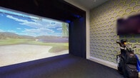 This single-level indoor golf simulator space is thoughtfully designed to deliver an immersive and functional golf experience. The centerpiece is a large, high-definition projection screen recessed within a dark-framed enclosure, displaying lifelike virtual golf courses—including scenic fairways, bunkers, and water features—powered by an ion simulator system. Subtle blue ambient lighting frames the screen bay, enhancing the visual atmosphere during play.

The surrounding walls are entirely clad in a striking honeycomb-patterned acoustic panel system, featuring gold geometric frames set against a grey upholstered backing. This distinctive design serves a dual purpose: it provides sound absorption to improve the acoustics of the room while acting as a bold visual statement throughout the space.

The floor transitions between dark grey carpet in the hitting area and a contrasting green artificial turf mat, marking the designated swing zone. A white bar-height counter along one wall is paired with a row of round-top adjustable stools, offering spectators a comfortable vantage point. A dark storage cabinet, wall-mounted display, and golf bag storage complete the setup. Entry is through a wide sliding wood-panel door, contributing to the room's clean, modern finish.