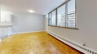 This thoughtfully laid-out unit offers a cohesive and functional living experience across four distinct spaces. The living room serves as the heart of the unit, showcasing a striking wall of large, floor-to-ceiling windows that flood the space with abundant natural light and frame views of the surrounding cityscape. Classic parquet hardwood flooring in a warm honey tone extends throughout the living areas, adding a timeless character to the interior. A baseboard heating system runs along the windowed wall, ensuring comfort year-round. The kitchen is efficiently designed with a galley-style layout, featuring white cabinetry with contemporary hardware, a granite-style countertop, a stainless steel sink, and white appliances including a refrigerator and electric range. Light tile flooring completes the clean, practical aesthetic. The bathroom is a standout feature, showcasing elegant marble-style wall panels surrounding a full soaking tub and shower combination, paired with a white vanity and modern chrome fixtures. A mirrored medicine cabinet adds both style and function. The hallway connects all areas seamlessly, with a mirrored sliding-door closet offering practical storage. Neutral grey walls throughout create a calm, versatile backdrop well-suited to a variety of interior styles.