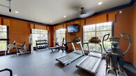 Located at 100 Stonecrest Blvd in Stonecrest, Georgia, this single-floor fitness center is a well-equipped facility designed to support a comprehensive workout routine. The space is defined by bold, warm-toned orange walls trimmed with deep navy crown molding, creating a vibrant and energizing atmosphere. Large multi-pane windows line the walls on multiple sides, flooding the interior with generous natural light and offering views of the surrounding greenery outdoors. A ceiling fan provides additional air circulation throughout the space.

The cardio area features multiple treadmills and an elliptical machine, all positioned to take advantage of the natural light from the large windows. A wall-mounted flat-screen television is centrally positioned for viewing during workouts. The strength training section is equally comprehensive, housing a Hoist multi-station cable machine, a weight bench with barbell rack, and a full dumbbell rack offering a wide range of free weights. Floor-to-ceiling mirrors on one wall enhance the sense of space and allow for proper form checks during exercise. The flooring throughout is durable rubber gym matting, suitable for both cardio and strength activities. Glass-paned doors provide access to an outdoor area, further connecting the interior to the natural surroundings.