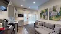 This well-appointed single-floor space combines a comfortable lounge area with a functional kitchenette in a clean, modern design. The open layout is anchored by warm wood-look flooring that transitions seamlessly into patterned carpet tiles, creating distinct zones within the room.

The kitchenette is outfitted with gray shaker-style cabinetry, marble-look countertops, a stainless steel sink, an over-the-range microwave, and a compact refrigerator. A water cooler and coffee station round out the amenities, making this area well-suited for everyday use. A wall-mounted flat-screen TV is positioned for easy viewing from the lounge.

The seating area features a generous gray sectional sofa with accent pillows, centered around a dark wood coffee table. Two coordinating abstract paintings in green and gray tones add a contemporary artistic touch to the neutral walls. Natural light enters through windows fitted with horizontal blinds and vertical blinds covering a sliding glass door, which provides access to the exterior. A wall-mounted mini-split HVAC unit ensures year-round climate comfort. Recessed lighting throughout keeps the space bright and inviting. The overall ambiance is clean, functional, and modern — well-suited as a breakroom, lounge, or common area.