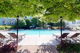 This thoughtfully designed outdoor amenity area offers a compelling blend of leisure and comfort within a lushly landscaped setting. At its centerpiece is a generously sized swimming pool, its clear blue water framed by a wide pool deck, classic lamp post lighting, and a canopy of mature trees that provide natural shade and a serene atmosphere.

Adjacent to the pool, a covered pavilion serves as the social hub of the space. Constructed with stacked stone accent walls and a solid roof with pendant lighting, the pavilion is both elegant and functional. A floor-to-ceiling stone fireplace with a mounted outdoor television anchors one end, creating a cozy gathering spot in any season. Stamped concrete flooring throughout the pavilion lends a refined, durable finish, while an assortment of seating—including rattan lounge chairs, dining tables with benches, and accent chairs—accommodates a variety of uses.

Beyond the pavilion, a brick-paved courtyard winds through well-maintained garden beds filled with ornamental grasses, flowering shrubs, and trimmed hedgerows. Wrought iron benches and shaded high-top dining tables with umbrellas are positioned throughout, offering additional outdoor seating options. A built-in grilling station provides a dedicated space for outdoor cooking and entertaining. The entire area is framed by the surrounding multi-story building, whose balconies overlook the courtyard, connecting the indoor and outdoor living experience seamlessly.