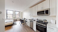 Located at 150 S Independence Mall W in Philadelphia's historic Old City neighborhood, this thoughtfully designed studio unit combines modern finishes with a practical, open-concept layout. The main living area features warm wood-tone flooring throughout, seamlessly connecting the kitchen, dining, and sleeping areas into one cohesive space. The kitchen is outfitted with white shaker-style cabinetry, light stone countertops, and a full suite of stainless steel appliances, including a range, over-the-range microwave, dishwasher, and refrigerator — providing everything needed for everyday cooking. A compact dining area with a round glass-top table and chairs sits adjacent to the kitchen, bathed in natural light from large black-framed windows that frame views of the surrounding Philadelphia cityscape. The sleeping area accommodates a full-size bed and benefits from dual windows overlooking the urban landscape below. The bathroom is finished with marble-look tile flooring, white subway tile walls, a glass-enclosed walk-in shower with a rainfall fixture, and a white vanity with ample storage. Clean, neutral tones and an efficient use of space define the overall aesthetic of this well-appointed unit.