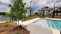 Located at 110 Frem Boustany Dr in Lafayette, Louisiana, this outdoor amenity space centers around a generously sized resort-style swimming pool that serves as the focal point of the community. The pool area is thoughtfully designed with a clean, modern farmhouse aesthetic, characterized by crisp white board-and-batten siding, dark-trimmed windows, and steeply pitched rooflines on the surrounding clubhouse and residential buildings.

The pool itself features a gradually sloping entry with in-water sun loungers, multiple staircase access points with stainless steel railings, and clear turquoise water. Flanking both sides of the pool are expansive concrete pool decks lined with chaise lounge chairs and low-profile tables, offering ample space for relaxation and socializing. Structured pergolas with slatted wood roofs anchor each corner of the deck, providing shaded retreats from the Louisiana sun.

Lush landscaping, including young trees and mulched planting beds, adds a natural warmth to the hardscaped environment. Tall pole-mounted light fixtures ensure the space is well-lit for evening use. A black iron perimeter fence provides both security and a refined visual boundary. The adjoining clubhouse building, with its floor-to-ceiling windows and direct pool access, creates a seamless indoor-outdoor connection, reinforcing the community's upscale, amenity-rich character.