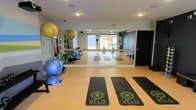 This single-floor fitness studio is thoughtfully designed to support a wide range of exercise disciplines. The space features a full-length mirrored wall that visually expands the room while allowing participants to monitor their form during workouts. A ballet barre runs along the mirrored wall, making the studio suitable for barre, yoga, and stretching sessions. The light wood-toned flooring provides a clean, durable surface underfoot, while recessed ceiling lighting ensures the space is well-lit throughout.

Branded "Vélo" yoga mats are laid out across the floor, indicating a dedicated group fitness environment. Along one side of the studio, an organized equipment rack holds stability balls in multiple sizes, resistance tools, and balance trainers such as BOSU balls, offering a comprehensive selection of fitness accessories. A window toward the back allows natural light to filter in, contributing to a bright and energizing atmosphere.

The studio's layout is open and flexible, accommodating group classes or individual training sessions with ease. Overall, the space presents a clean, professional, and well-equipped environment suited for diverse fitness activities.