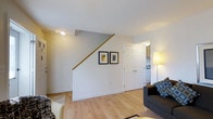 This two-story, two-bedroom, one-bathroom home offers a functional and well-organized layout across two levels. The main living area is visible upon entry, featuring light hardwood-style flooring that extends throughout the space, contributing to a bright and cohesive aesthetic. The living room accommodates comfortable seating and is illuminated by both ambient and accent lighting, creating a warm atmosphere.

A staircase with a natural wood handrail rises along one wall, providing access to the upper floor while serving as a subtle architectural detail in the main living space. Framed artwork adorns the wall alongside the staircase, adding a finishing touch to the area. Neutral white walls and white trim throughout keep the interior feeling clean and airy.

Adjacent to the living area, a doorway leads toward the kitchen, which features cabinetry and appliances visible from the main room. The overall flow between spaces is straightforward and practical. The two-level design allows for a natural separation between living and sleeping areas, offering residents a degree of privacy and organization within the home.