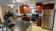 This kitchen is thoughtfully designed with a cohesive aesthetic that balances practicality and visual appeal. The cabinetry, finished in a warm cherry wood tone, extends from floor to ceiling and includes glass-front upper cabinet doors that add a refined touch. Some cabinets feature decorative details and are topped with tasteful greenery, lending the space a lived-in, welcoming feel.

Dark laminate countertops provide a striking contrast against the rich wood tones and run along an L-shaped layout, offering generous prep and workspace. The kitchen is anchored by a peninsula that extends the counter space further, accommodating casual seating or additional workspace.

Stainless steel appliances, including a built-in microwave, an electric range with oven, and a full-size refrigerator, are seamlessly integrated into the cabinetry. A double-basin stainless steel sink sits beneath the countertop with easy access to storage below.

Track lighting installed on the ceiling provides bright, directional illumination throughout the space. Light-toned tile flooring completes the look, adding brightness and easy maintenance underfoot. The overall ambiance is warm, functional, and polished.