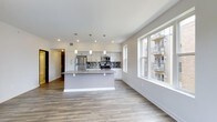 This 2-bedroom, 2-bathroom residence is thoughtfully designed with a single-floor, open-concept layout that promotes a seamless flow between living and kitchen areas. The kitchen serves as a central focal point, anchored by a sizeable island with a sleek gray finish that doubles as a breakfast bar and prep space. White shaker-style cabinetry lines the walls, complemented by stainless steel appliances, including a refrigerator, microwave, and range. A bold mosaic tile backsplash adds a striking design element, while pendant lighting above the island provides both task lighting and visual interest. Recessed ceiling lights further illuminate the space throughout. Wide-plank wood-style flooring runs continuously across the living and kitchen areas, lending warmth and cohesion to the contemporary aesthetic. Large double-hung windows along the living area wall invite abundant natural light and offer views of the surrounding urban streetscape. Clean white walls and modern trim throughout contribute to a crisp, airy atmosphere. A hallway leads to the two bedrooms and bathrooms, providing a clear separation between living and private spaces.