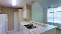 This two-story property presents a thoughtfully arranged floor plan that maximizes functionality across both levels. The overhead layout reveals a well-organized main floor with distinct zones for living, cooking, and bathing, connected by a staircase that leads to the upper level. The kitchen is a notable feature, equipped with white cabinetry providing ample storage and a double-basin stainless steel sink set into white countertops. The angled ceiling above the kitchen adds architectural character, contributing to a sense of varied ceiling height throughout the space. Light tiles cover the kitchen floor, enhancing the brightness of the room, which also benefits from a window that allows natural light to filter in. A pass-through counter connects the kitchen to an adjacent area, supporting an open and communicative layout. The bathroom areas, visible in the floor plan, are efficiently positioned for convenient access. Warm-toned flooring appears throughout much of the main level, complementing the neutral wall palette. The staircase, visible in the lower portion of the floor plan, provides a clear transition between the two floors, emphasizing the vertical dimension of this multi-level space.