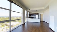 This single-floor unit is situated on an upper level of a high-rise building, offering dramatic panoramic views of the surrounding cityscape, lush tree canopy, and a expansive lake stretching to the horizon. A private balcony with a sleek metal railing provides direct access to these views, making it an ideal vantage point to take in the skyline, which includes a notable domed capitol building in the distance.

Inside, the open-concept layout is defined by large floor-to-ceiling windows that flood the living area with abundant natural light while framing the stunning outdoor scenery. Rich, dark hardwood flooring runs throughout, lending warmth and contrast to the bright, neutral-toned walls and ceilings.

The kitchen is centrally positioned within the open living space, featuring dark wood cabinetry, dark granite or stone countertops, and stainless steel appliances including a refrigerator, microwave, and range. Pendant lighting above the kitchen island adds a refined, contemporary touch.

Together, the elevated position, expansive glazing, and modern finishes create a polished, light-filled living environment with a strong connection to the urban landscape beyond.