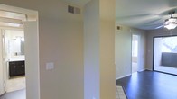 This single-floor space offers a clean, straightforward layout designed for functional everyday living. The interiors are finished in a warm neutral palette, with smooth walls painted in soft beige and green-tinted tones that lend a calm, understated ambiance throughout. The flooring transitions between dark hardwood and light ceramic tile, providing visual variety while delineating different areas of the space. Ceiling-mounted HVAC vents are present in multiple areas, indicating a central air system that serves the full layout. Structural columns and partial walls create gentle separations between spaces without fully closing off the floor plan, contributing to an open and connected feel. Natural light is drawn in through windows visible toward the end of the hallway, brightening the interior and adding a sense of airiness. White baseboards run along the walls, adding a crisp finishing detail to the overall design. The space presents a well-maintained, move-in ready environment with a no-frills aesthetic that emphasizes practicality and ease of use. Its single-level design ensures seamless accessibility throughout, making it a straightforward and efficient space to navigate.