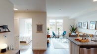 This single-floor unit showcases a thoughtfully designed open-concept layout that seamlessly connects the kitchen, dining, and living areas. The kitchen is outfitted with light wood-grain cabinetry extending to the ceiling, stainless steel appliances including a refrigerator, range, and over-the-range microwave, a white subway tile backsplash, and a peninsula with a dark quartz countertop that doubles as a breakfast bar. The living and dining area benefits from large windows that draw in generous natural light and frame outdoor views of the surrounding landscape. The space accommodates a casual dining setup alongside a comfortable lounge area with a glass coffee table. The bedroom is a standout with a bold teal accent wall, a large window offering outdoor views, carpeted flooring, and ample wall space for artwork. The bathroom features a combination tub and shower, wood-grain vanity cabinetry, a dark quartz countertop with an undermount sink, and a large mirror. Warm hardwood-style flooring runs throughout the main living areas, contributing to the unit's cohesive, contemporary aesthetic.