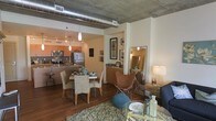 This stylish urban loft presents a thoughtfully designed open-concept floor plan that seamlessly blends the kitchen, dining, and living areas into a cohesive, flowing space. The industrial character of the unit is immediately evident through the exposed concrete ceiling and prominent ductwork, lending an authentic loft aesthetic that is both contemporary and inviting. Rich hardwood floors run throughout, anchoring the space with warmth and continuity. The kitchen features stainless steel appliances, sleek cabinetry in warm orange-toned wood, and a peninsula that doubles as additional prep and seating space. The dining area accommodates a full table with mixed seating, including woven and upholstered chairs, creating a casual yet refined atmosphere. Adjacent to the dining space, a dedicated living area is defined by a plush area rug and comfortable seating, while a compact accent console with decorative accessories adds visual interest. A mirror near the living area reflects light and adds a sense of depth to the room. Pendant lighting throughout the space provides focused illumination, complementing the overall modern industrial design. The single-floor layout ensures easy navigation and a unified ambiance from room to room.