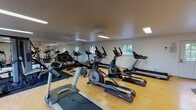 Located at 259 Colonnade Dr in Charlottesville, Virginia, this fitness center offers a well-organized, single-floor workout facility designed to support a range of exercise routines. The space features light wood-tone flooring throughout, complemented by clean white walls and a bright, functional ceiling with recessed and fluorescent lighting that ensures even illumination across the entire area. Multiple windows are positioned along the walls, inviting natural light and providing pleasant views of the surrounding greenery, creating an open and energizing atmosphere. A full-length mirrored wall spans one side of the room, enhancing the sense of space and allowing users to monitor their form during workouts. The cardio section is well-stocked with multiple treadmills, elliptical trainers, and stationary bikes, accommodating several users simultaneously. A dedicated strength training area features a multi-station cable machine, a rowing machine, and a weight bench, providing options for full-body resistance training. Rubber floor mats are placed beneath the equipment to protect the flooring and reduce noise. The layout is thoughtfully arranged to maximize circulation and usability, with open floor space available for stretching or additional exercises. The fitness center is accessible via a dedicated entry door and offers a clean, practical environment suited for everyday fitness use.