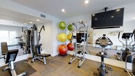 This single-floor fitness center is thoughtfully designed to support a well-rounded workout routine. The space features a clean, modern aesthetic with white walls, recessed lighting, and neutral carpet tile flooring that creates a functional yet inviting environment. Large mirrors line the walls, enhancing the sense of space and allowing users to monitor their form during exercise. The gym is equipped with a multi-function cable machine and a power tower for strength training, complemented by a selection of exercise balls in varying sizes for flexibility and core work. A stationary bike provides cardio options, and a wall-mounted flat-screen television adds an entertainment element to workouts. The recessed ceiling lights provide bright, even illumination throughout the space, ensuring excellent visibility at all times. The layout is efficiently organized, allowing for ease of movement between equipment. Ventilation is supported by ceiling-mounted air vents, maintaining a comfortable climate during use. Overall, this fitness center offers a compact yet comprehensive facility suitable for a variety of workout styles and fitness levels.