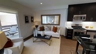 This single-floor space offers a well-designed open-concept layout that seamlessly integrates the living and kitchen areas into one cohesive, functional environment. The living area is centered around a clean-lined white sofa accented with warm earth-toned throw pillows, complemented by a sleek dark coffee table with gold-toned legs resting atop a textured woven area rug. A large framed mirror above the sofa enhances the sense of depth and light, while a pair of framed black-and-white art pieces adds a contemporary touch to the neutral walls. Natural light filters through a window behind the sofa, brightening the space. The kitchen, positioned adjacent to the living area, features dark cabinetry with a rich, espresso-toned finish, stainless steel appliances including a range and microwave, and a clean white tile backsplash that contrasts elegantly with the dark finishes. Warm wood-look flooring runs throughout the space, providing continuity and a grounded, inviting feel. Recessed lighting overhead ensures consistent illumination across the open floor plan. The overall ambiance is modern and refined, with a neutral color palette punctuated by warm accent tones that create a welcoming and sophisticated atmosphere.