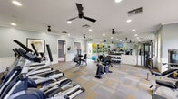 Located at 2437 Promenade Blvd in Montgomery, Alabama, this single-story fitness facility offers a comprehensive and thoughtfully designed workout environment. The main exercise area is spacious and open, finished with a distinctive patterned carpet in neutral tones of gray, tan, and yellow that adds visual warmth to the space. Light blue-gray walls with classic white wainscoting paneling create a clean, polished aesthetic throughout.

The fitness floor is well-appointed with a wide variety of equipment, including treadmills, stationary bikes, stair climbers, and multiple strength-training machines. A full rack of dumbbells and an assortment of exercise balls in vibrant colors add versatility for varied workout routines. A large floor-to-ceiling mirror runs along one wall, visually expanding the space and providing useful feedback during exercise. Multiple black ceiling fans and recessed lighting ensure the room remains comfortable and well-lit.

Natural light filters in through several windows, lending an airy, inviting feel to the space. An adjoining playroom connects directly to the fitness area, featuring the same carpet, large windows with tailored valances, and a colorful world map mural — making it a welcoming space for younger visitors.
